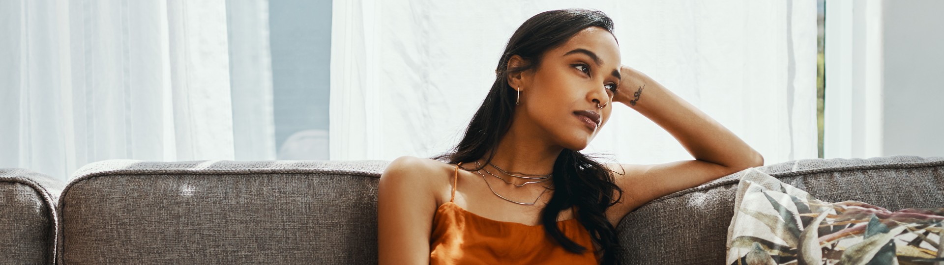 Woman with long dark hair and wearing an orange top leaning back on a couch and gazing to the side